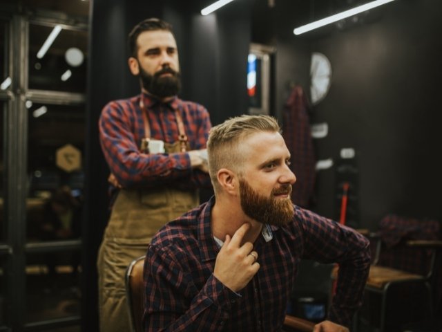 Profissional de barbearia em é com braços cruzados enquanto seu cliente avalia o serviço que ele fez  