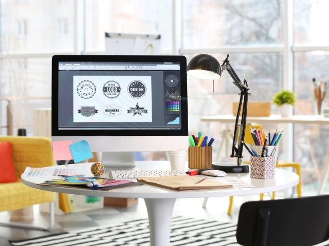 Curso de design gráfico online - imagem de um computador com uma tela aberta, vários papeis e lápis sobre a mesa