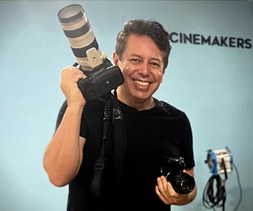 Fotografia do Professor Fábio Medeiros sorrindo segurando uma camera fotografica na mão esquerda e ooutra na mão direita.